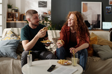 Happy young couple sitting on couch in living room and discussing movie while eating hamburgers and...