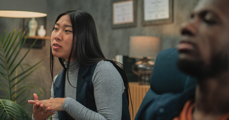 Multiethnic young couple on counseling session with psychologist. Asian young woman talks about relationship conflict. In the foreground, unfocused unhappy African American boyfriend. Couples therapy.