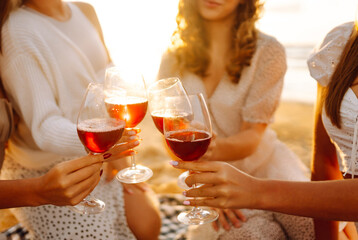 Four friends celebrate together at the beach, raising their glasses of red wine. The sun sets in the background, creating a warm and joyful atmosphere. Hen party.