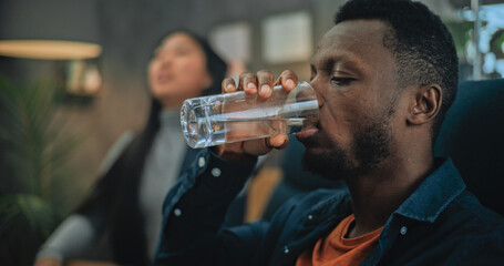 Close up of annoyed African American man listening his girlfriend, drinking water during couples therapy. In the background Asian woman discusses relationship with therapist on counseling session.