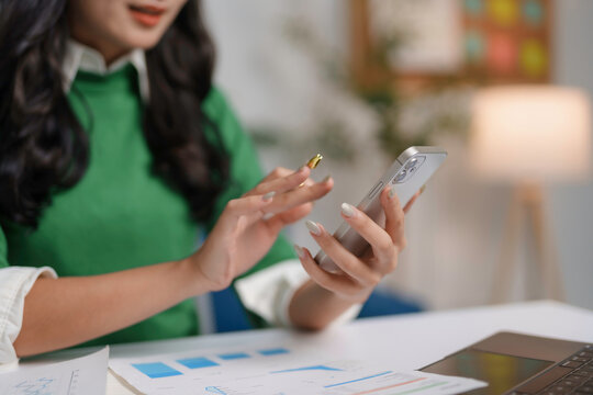 Businesswoman is using a smartphone and stylus pen while reviewing financial charts and graphs at her desk - Powered by Adobe