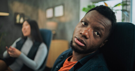 Close up of tired African American man listening his girlfriend, drinking water during couples therapy. In the background Asian woman discusses relationship with psychologist on counseling session.