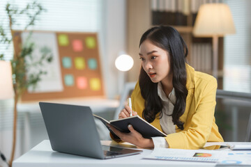 Fototapeta premium Young businesswoman is taking notes in her planner while working on her laptop in her bright and modern office