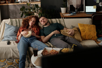 Young relaxed couple with popcorn napping on comfortable couch in front of table with junk food and...