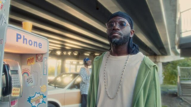 Confident Black man in durag and casual outfit posing at camera next to sticker-covered payphone, his friend listening to music by retro car under urban overpass. Video portrait, zoom shot