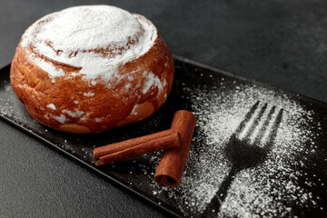 Round, delicious bun with cinnamon and powdered sugar on a black plate on a gray background