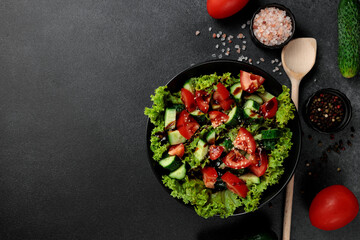 Vegetable salad of tomatoes, cucumbers, onions, lettuce dressed with aromatic dressing in a black plate on a gray background