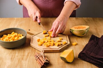 Woman in brown apron and shirt cutting pumpkin into cubes on wooden board. Cooking pumpkin dishes.