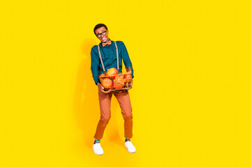 Full size photo of pretty young male hold shopping basket pumpkins heavy suffer wear trendy suit outfit isolated on yellow color background