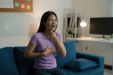 Obraz premium Young woman suffering from sore throat touching her neck with painful expression, sitting on sofa at home