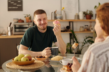 Young man looking at his girlfriend with smile while sitting by kitchen table in front of her and having donut with tea or coffee