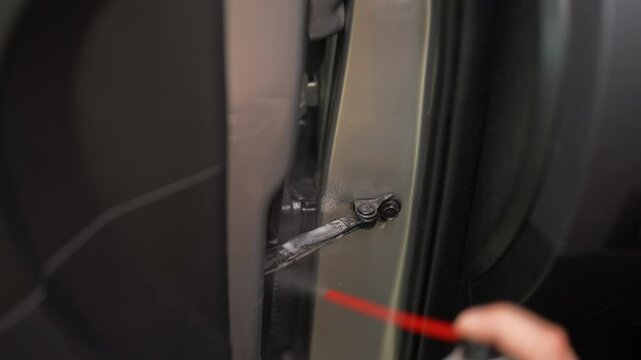 Mechanic male applying anti-corrosion grease to hinges of car door, ensuring smooth operation and preventing rust. Closeup of detailer greasing auto door hinge for smooth operation in workshop.