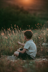 A boy in nature, about seven years old, rear view, with blond hair, wearing a beige T-shirt and black shorts, sitting on the grass against the background of the sunset. 