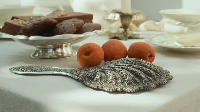 Close up view of ornate silver hand mirror lying on table near fresh apricots, tray of sweets and porcelain teacup outdoors on sunny day