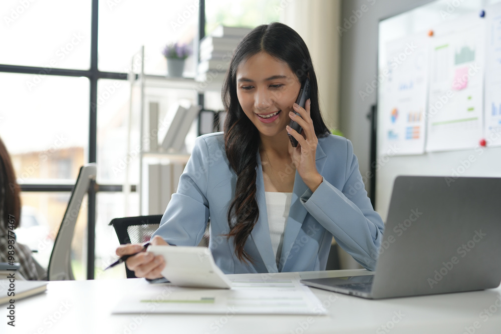 Aufkleber smiling businesswoman is using a calculator and taking notes while having a business call