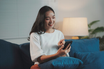 Young woman is using a smartphone and smiling while relaxing on a blue sofa at home at night