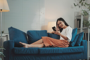 Obraz premium Young woman is lying comfortably on a sofa, browsing social media on her phone and smiling