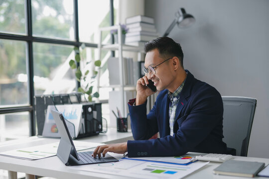 Asian businessman is smiling while working on a tablet and talking on the phone in his bright office