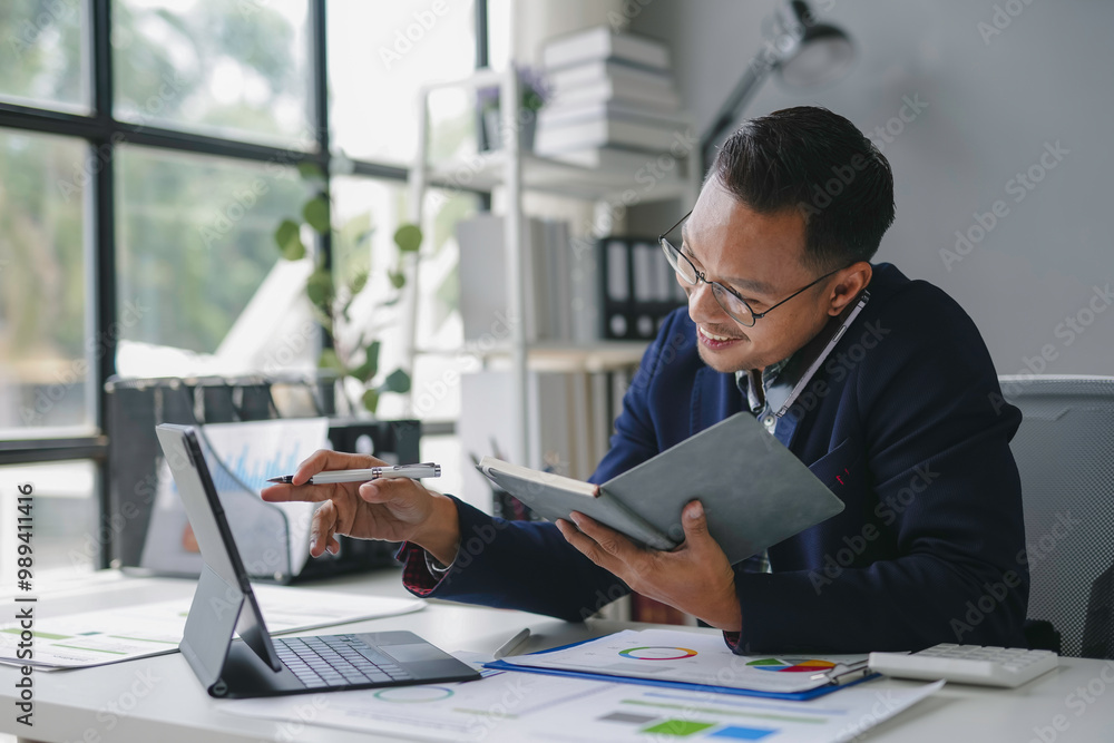 Poster businessman is working in the office, smiling while comparing data on his tablet and taking notes in