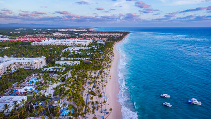Aerial view of Bavaro Beach, Punta Cana, Dominican Republic, captured at sunrise. Golden sandy coastline, lined with palm trees and resorts, meets the calm blue ocean with anchored boats (April 2022).