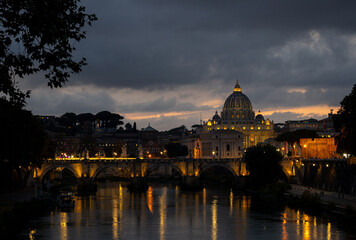 Fototapeta premium Evening view of St. Peter's Basilica across the Tiber River with illuminated buildings and dramatic clouds in Rome