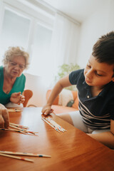 A young boy and his grandmother enjoy a pick-up sticks game in a warm and cozy living room setting,...
