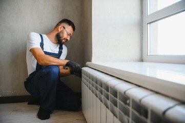 A plumber in a blue jumpsuit is fixing a radiator. With an adjustable wrench, he tightens the tap....