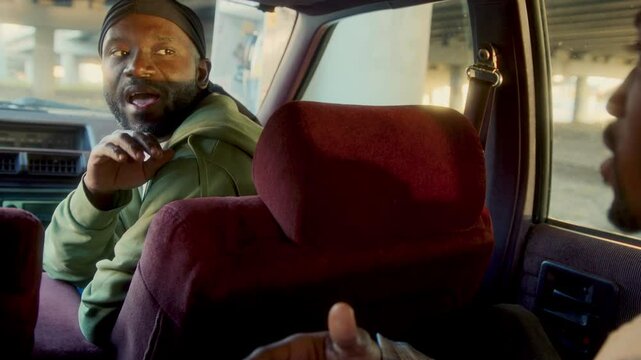 Young Black man wearing durag and casual outfit chatting with friend while sitting in backseat of vintage car under urban overpass. Rack focus