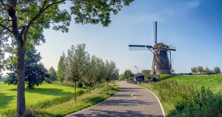 A winding road leads to a traditional windmill set against a clear blue sky. Lush green fields and trees line the sides of the road, creating a picturesque rural scene. © john