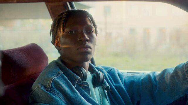 Young Black man with dreadlocks wearing denim jacket sitting on drivers seat of a car, listening to rap music, bobbing his head to the beat and looking seriously into camera. Video portrait