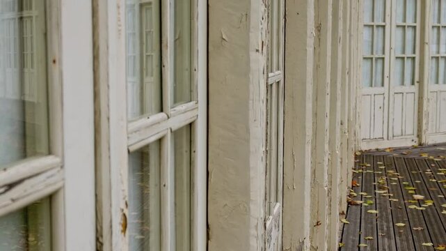 Old aged cracked wooden windows of beige manor veranda. Close up historical outbuildings 4K. Fallen leaves on terrace floor. Vintage shabby building. Cultural heritage object. XIX century architecture
