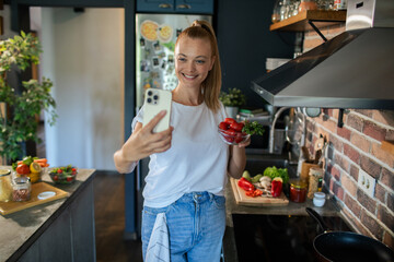 Smiling young woman cooking and filming with smartphone in modern kitchen