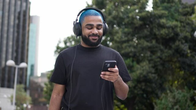 Cheerful Bearded Guy Wearing Headphones Walking Outdoors Using Smartphone Music New App in Modern City on Summer Day. Urban street. People and music concept. Stylish millennial male enjoying free time