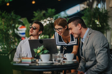 Business professionals gather outdoors with a laptop, collaborating in a relaxed cafe environment, surrounded by greenery and enjoying coffee.