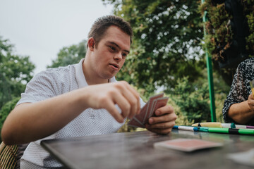 A young man with Down syndrome enjoys a card game outdoors, showcasing leisure and inclusion. He's...