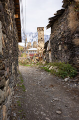 A narrow stone alleyway leading to historic stone towers in Ushguli, Georgia, with snow-capped mountains in the background. The image emphasizes the rustic charm and ancient architecture of the Svanet
