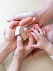 A baby hand holds a small wooden toy house together with the hands of its parents.