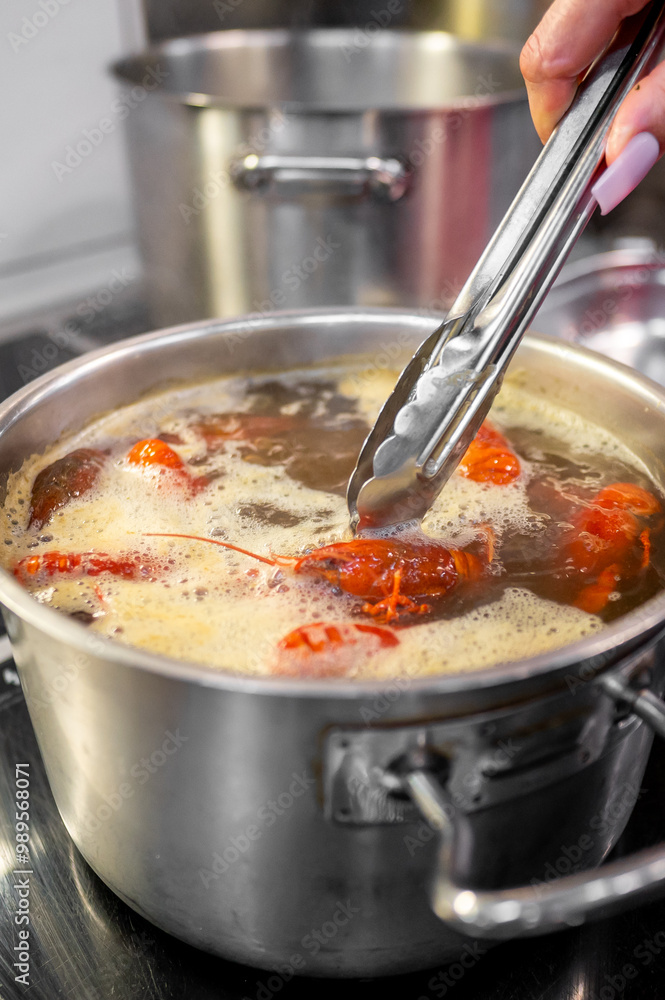 Wall mural close-up of a hand using tongs to cook soup in stainless steel pot on a stove. steam rises from the 