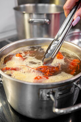 close-up of a hand using tongs to cook soup in stainless steel pot on a stove. Steam rises from the...