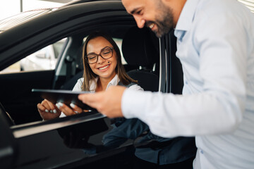 Woman in car talking with man holding tablet outside vehicle in a car showroom