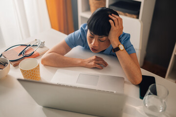 Weary asian woman medical professional in blue scrubs holding head by laptop