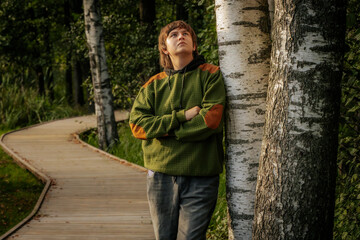 A young man stands with arms crossed, leaning against a birch tree, gazing upwards in thought. The scene is set along a wooden path in a serene forest.