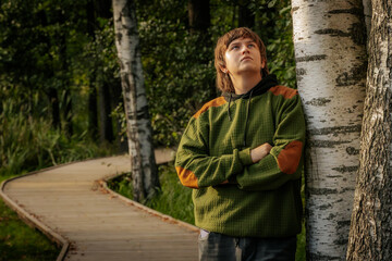 A young man stands with arms crossed, leaning against a birch tree, gazing upwards in thought. The scene is set along a wooden path in a serene forest.