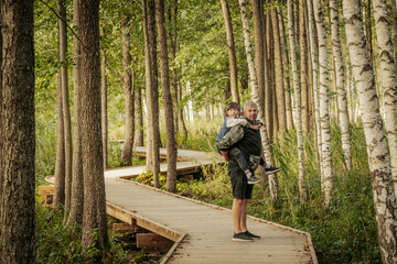 A smiling father gives his young son a piggyback ride on a wooden path through a forest. Both are...