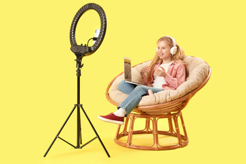 Teenage blogger in headphones with modern laptop sitting in armchair and recording video on yellow...