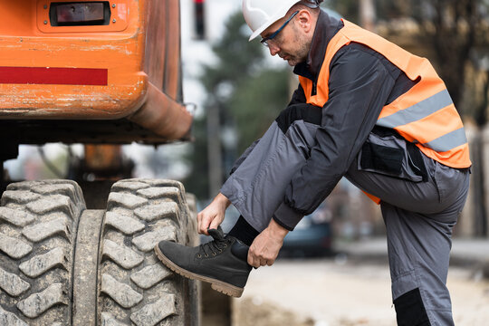 Construction worker adjusting boot while preparing for work near heavy machinery at a busy urban site in the afternoon