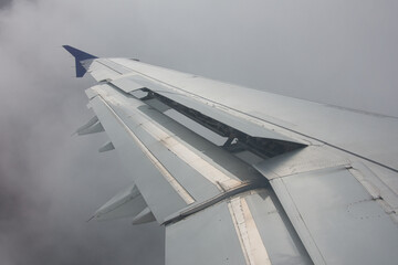 Close-up of an airplane wing with extended flaps in dense fog, air traffic above the clouds.