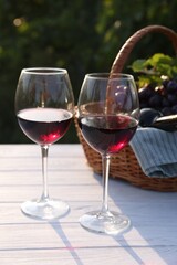Tasty wine in glasses, basket with bottle and grapes on white wooden table outdoors