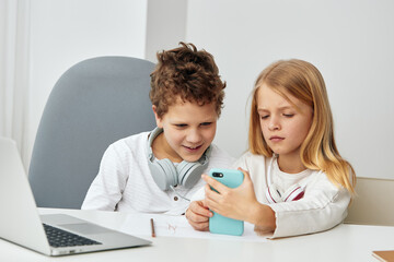 Happy children studying together at home A boy and a girl are sitting at a table in their cozy living room, using laptops and headphones for online learning With smiles on their faces, they are