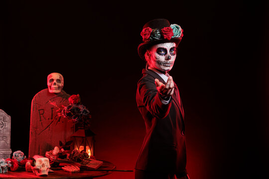 Portrait of woman doing enticing hand invitation in graveyard during Day of the Dead. Mysterious lady does provocative luring gesturing, surrounded by frightening funeral items during Halloween event